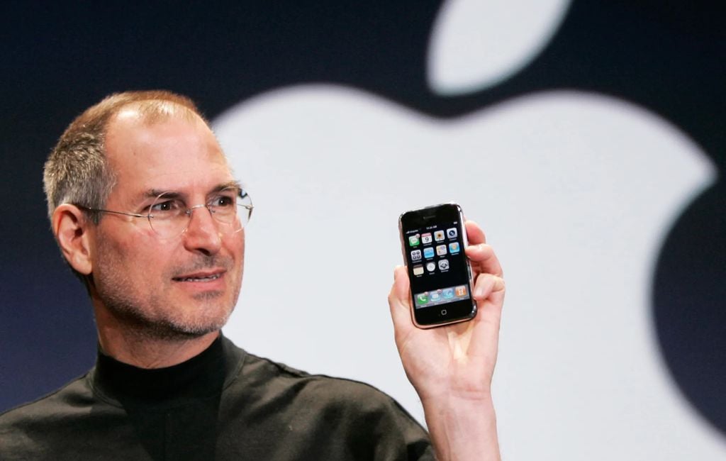 Steve Jobs apresenta um iPhone em suas mãos enquanto a logo da Apple aparece ao fundo