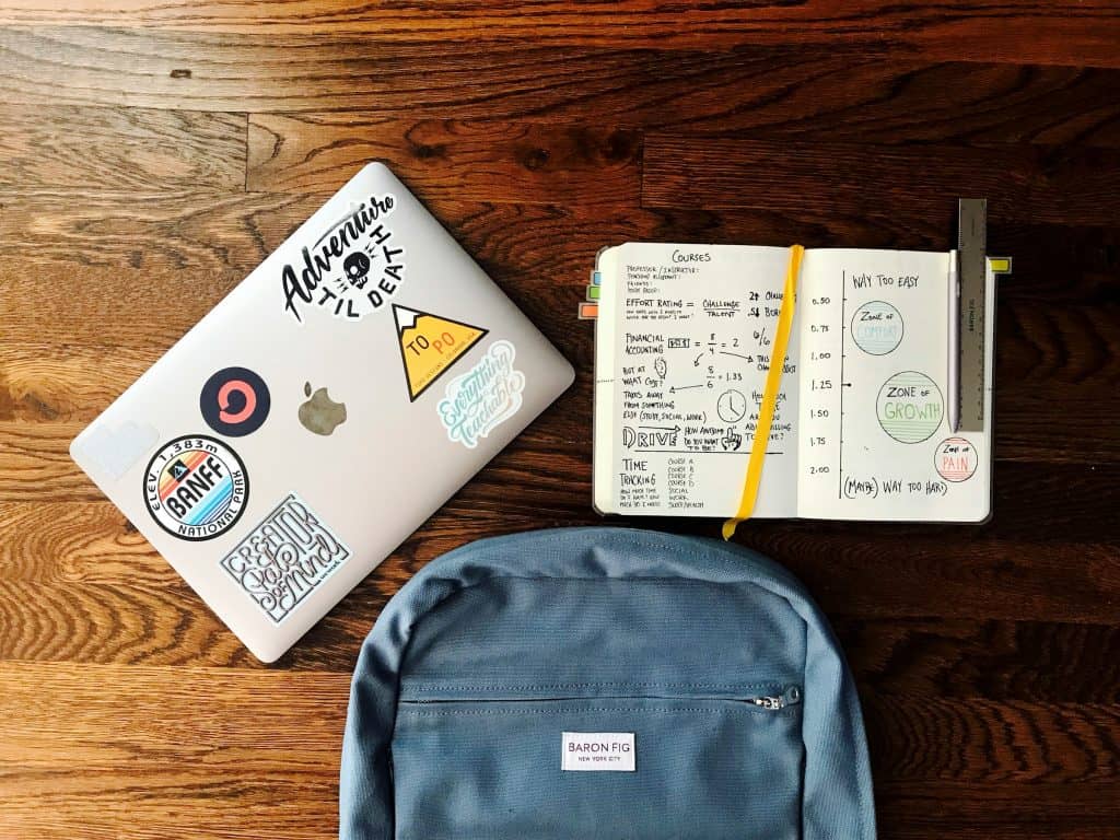 imagem mostra três objetos em cima de uma mesa marrom de madeira: uma mochila de escola, a parte de cima de um laptop fechado e um caderno aberto com várias anotações à mostra