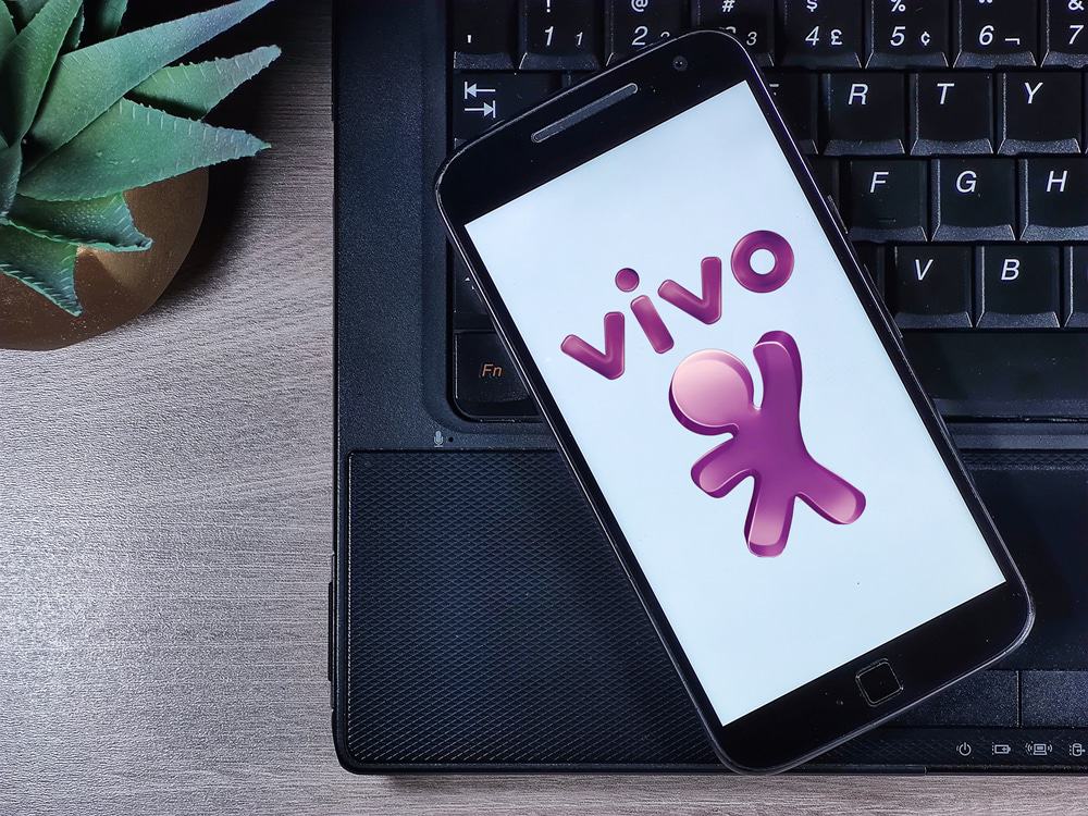 Em cima de uma superfície de madeira, aparece um teclado de notebook e, em cima dele, um smartphone com o logotipo da operadora Vivo na tela.