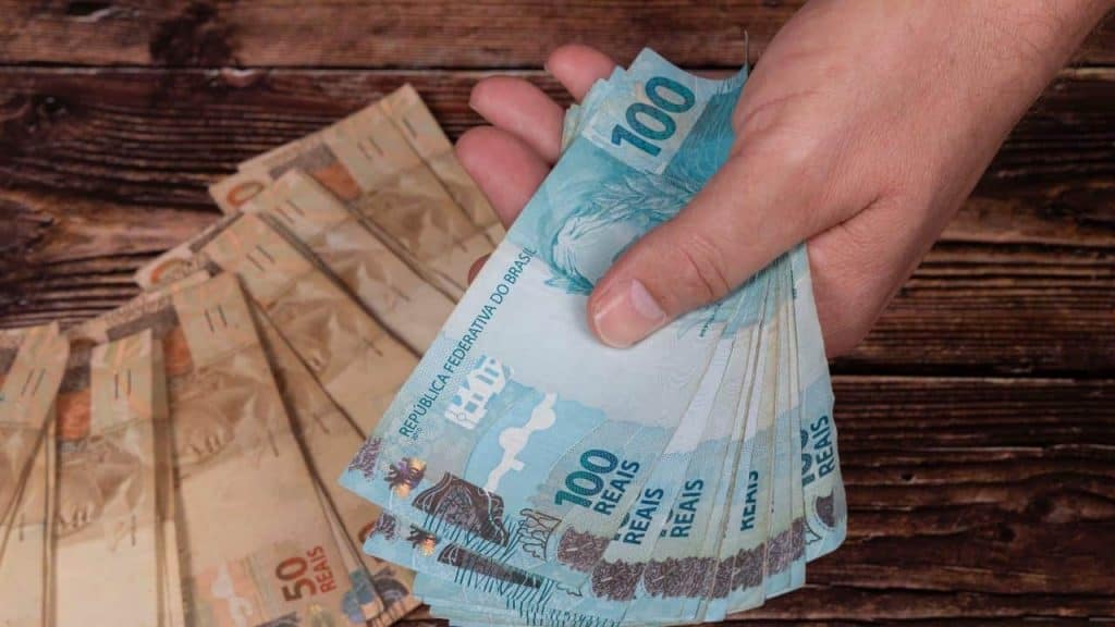 Notas de R$ 100 na mão de uma pessoa; abaixo, em uma mesa, notas de R$ 50